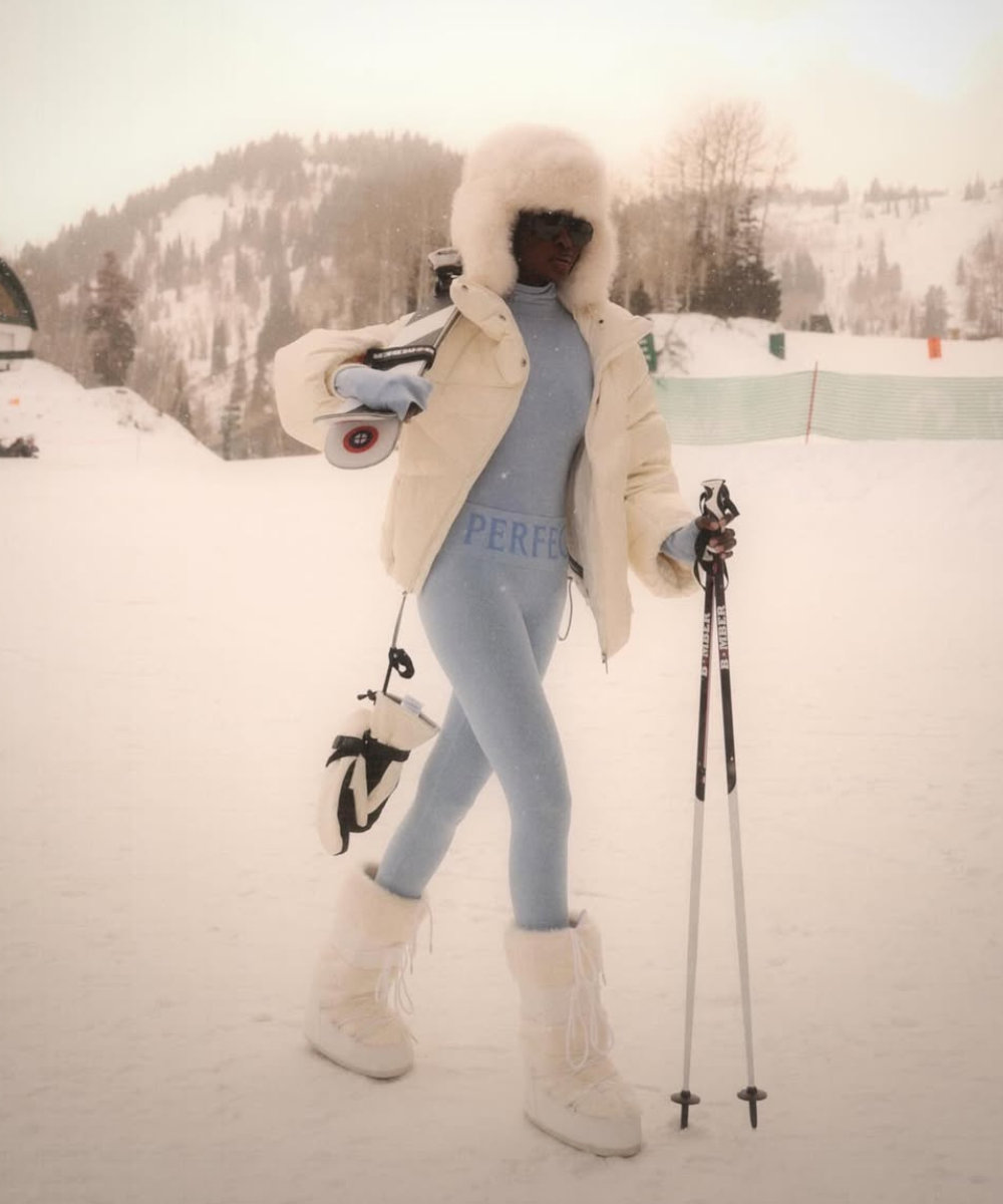 Pessoa em pista de esqui com look estiloso. Usa traje azul, botas brancas e casaco peludo. Montanha ao fundo.
