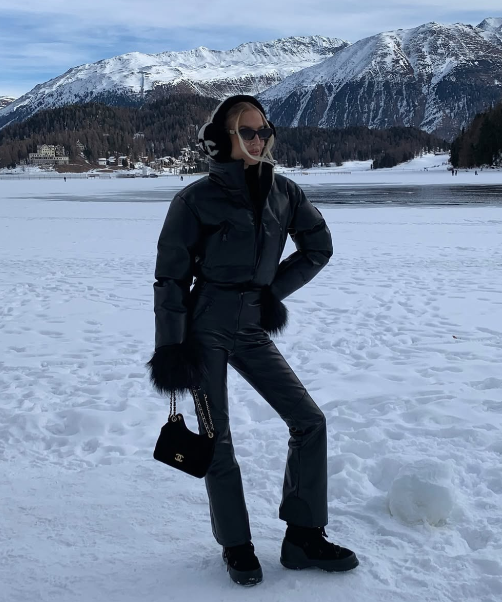 Pessoa em roupa preta elegante com acessórios de pele na neve, montanhas ao fundo, exibindo looks estilosos para esquiar.