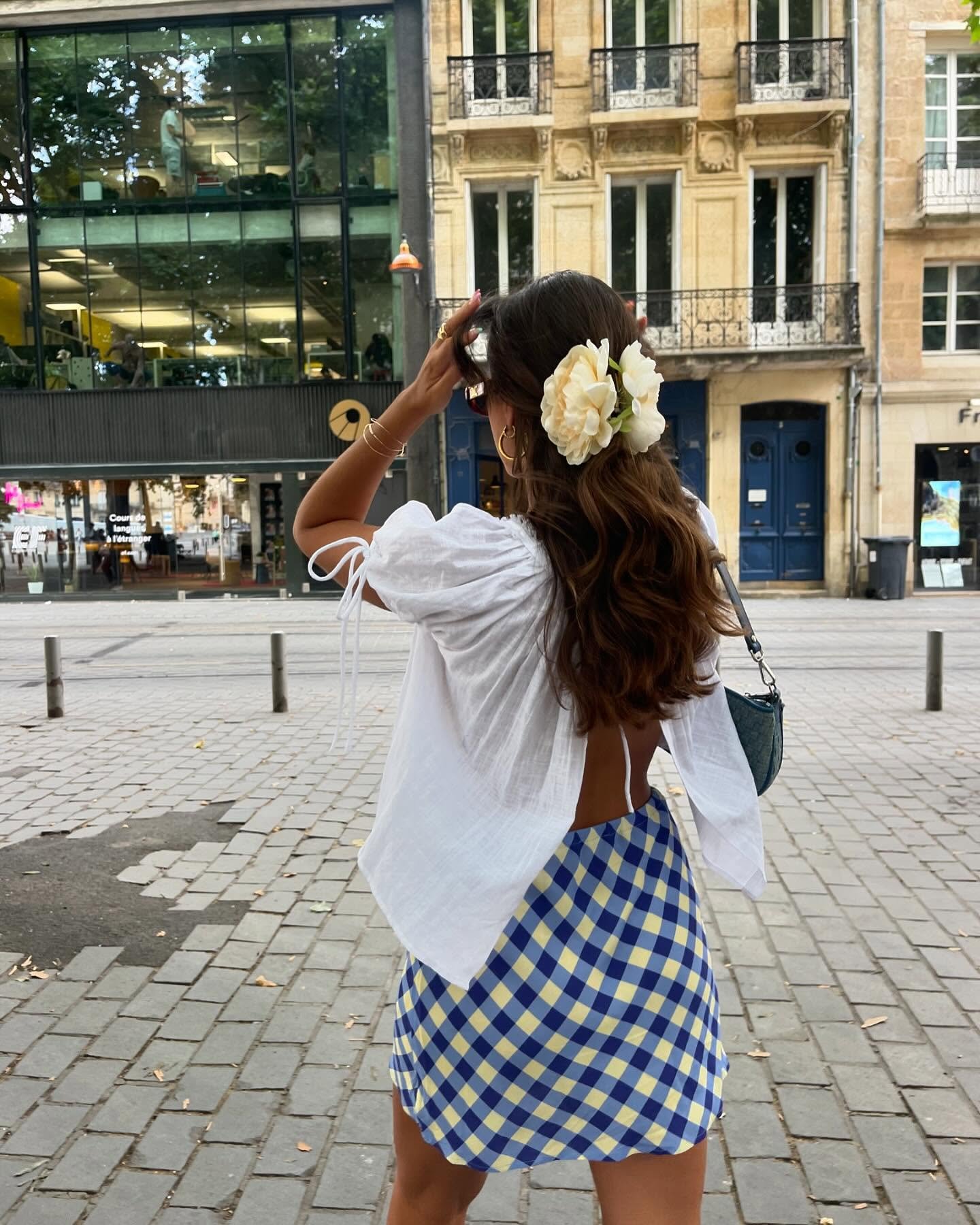 Mulher com vestido xadrez azul e branco de costas numa rua, com flores no cabelo.