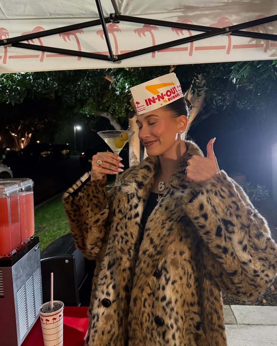 Pessoa com casaco de pele e chapéu de papel, segurando drink e fazendo sinal de positivo. Loud luxury
