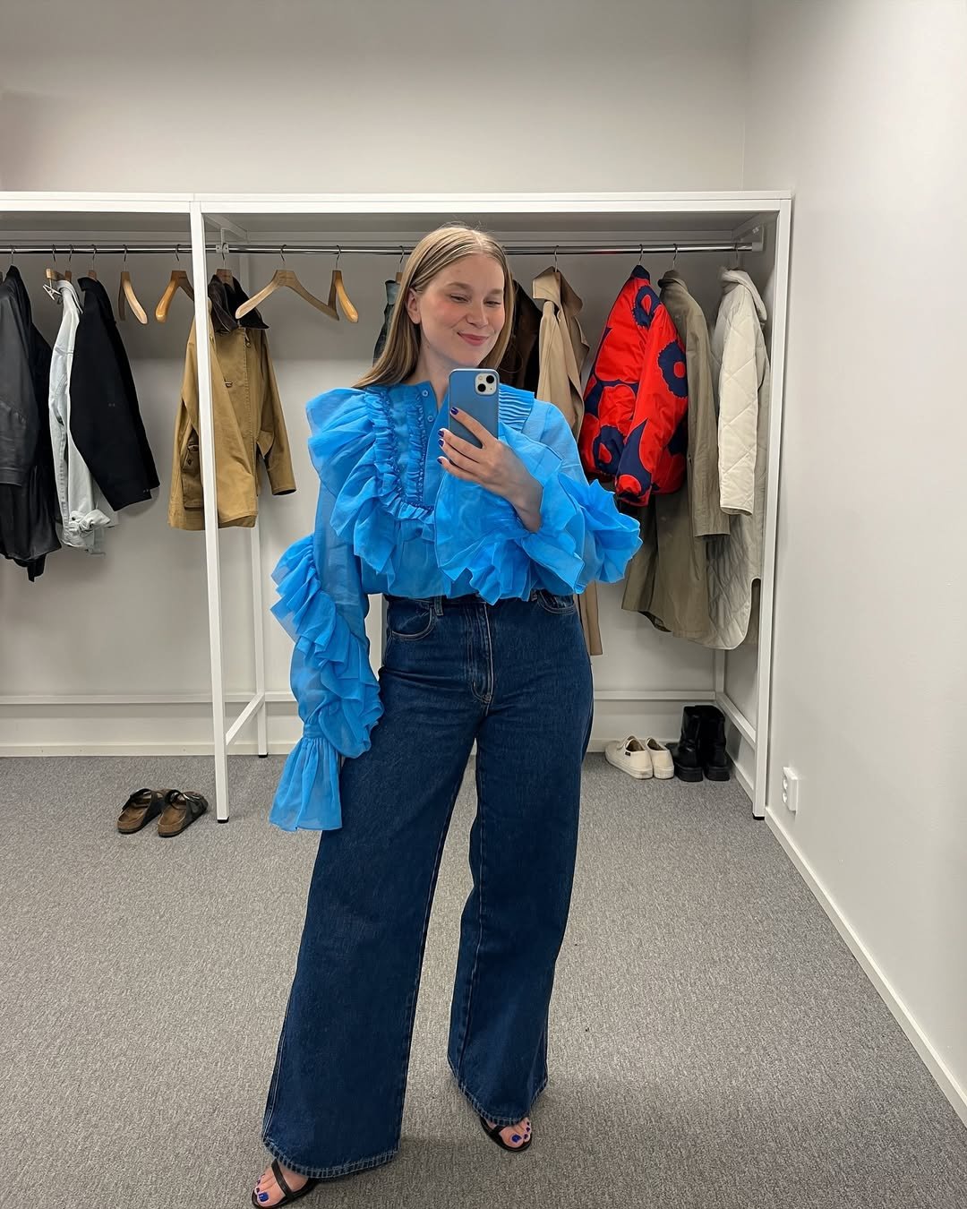 Pessoa vestindo blusa azul vibrante com babados e calça jeans em um closet repleto de roupas elegantes e sapatos. Loud luxury
