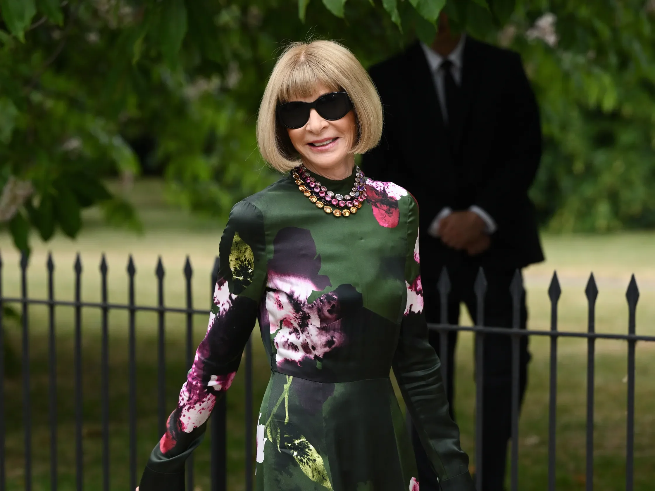 Uma pessoa com vestido floral verde e óculos escuros sorri ao ar livre, expressão de luxo discreto.