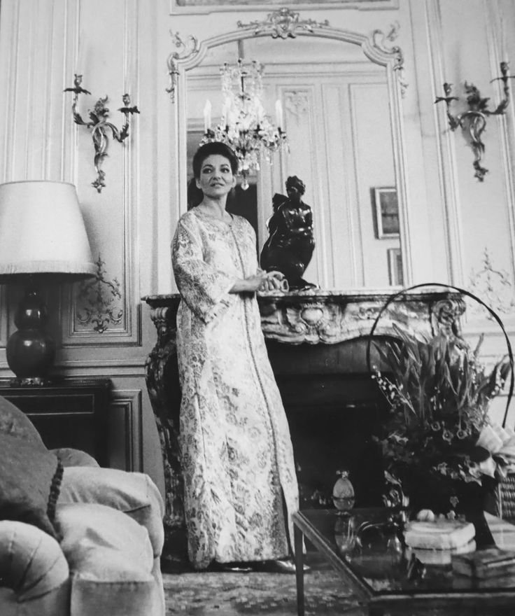 Maria Callas em pé em sala elegante com candelabro e escultura na lareira, vestindo robe decorativo.