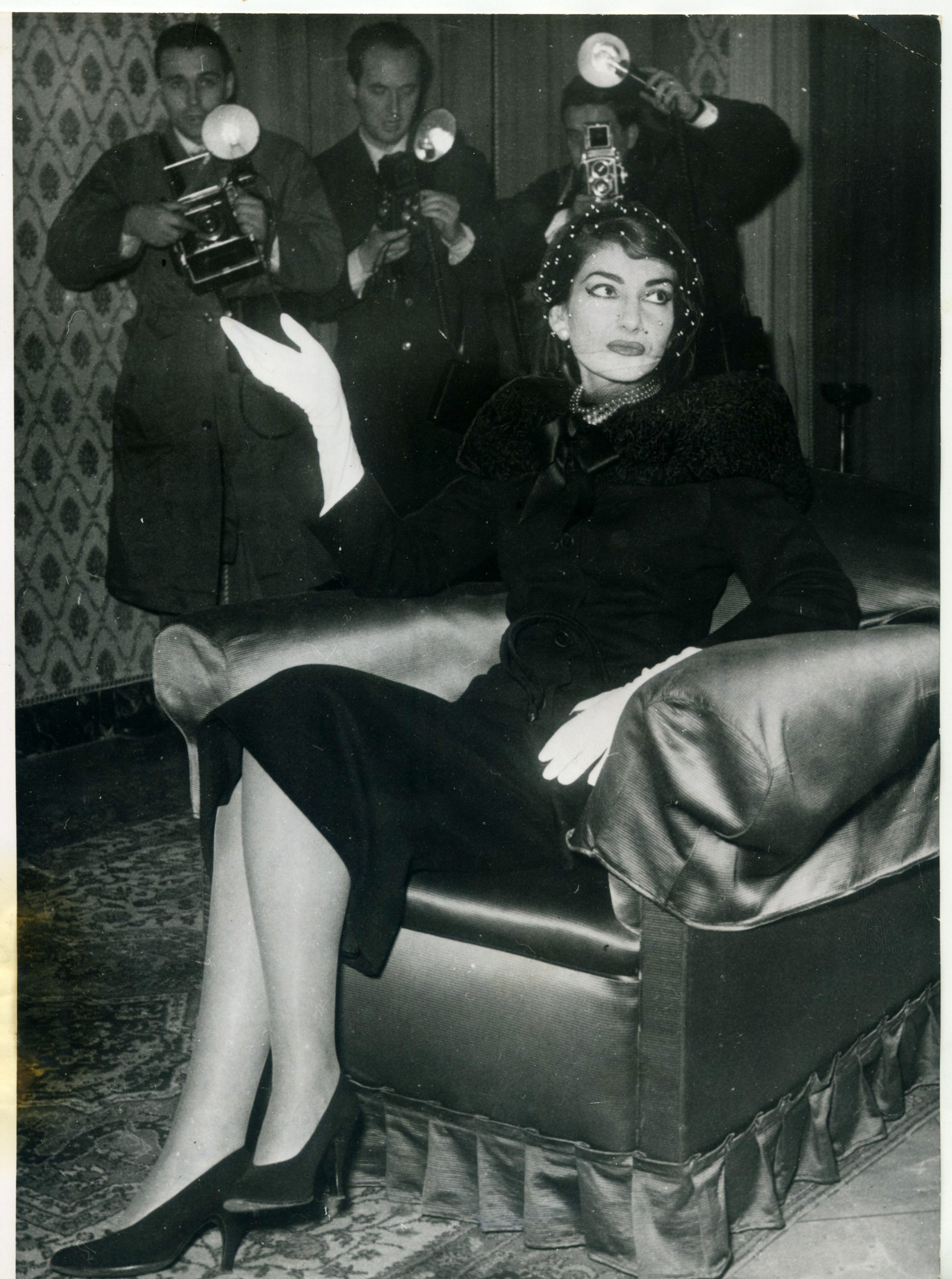 Maria Callas um sofá, cercada por fotógrafos com câmeras antigas.