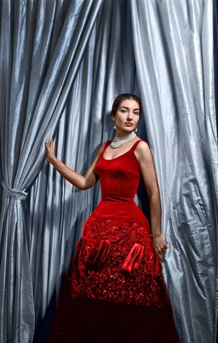 Maria Callas em vestido vermelho elegante, entre cortinas de cetim prateadas.