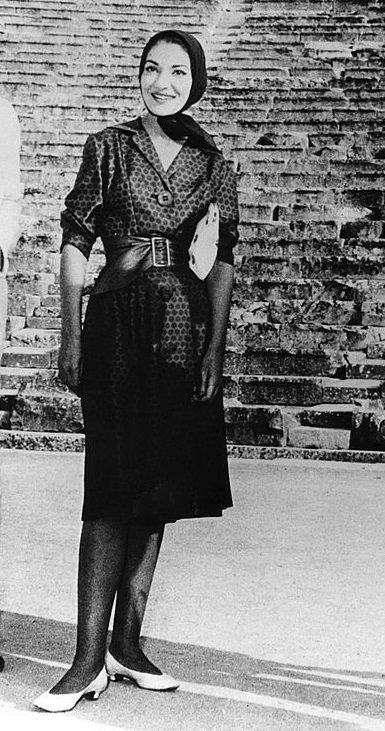 Maria Callas com vestido estampado e lenço na cabeça está em frente a degraus de pedra.