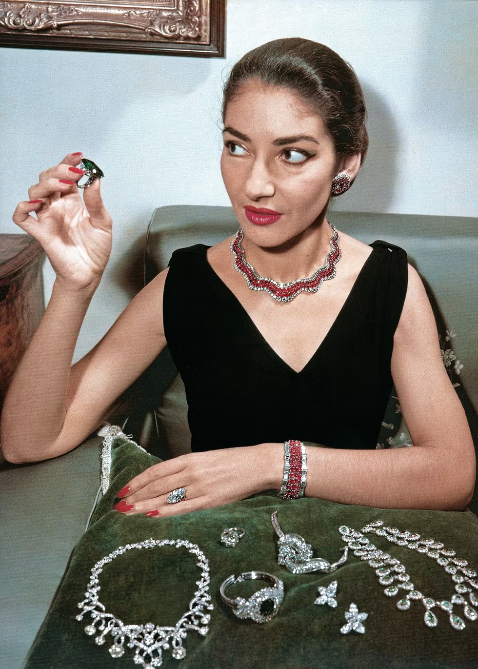 Maria Callas está segurando uma joia, cercada por colares e braceletes reluzentes.