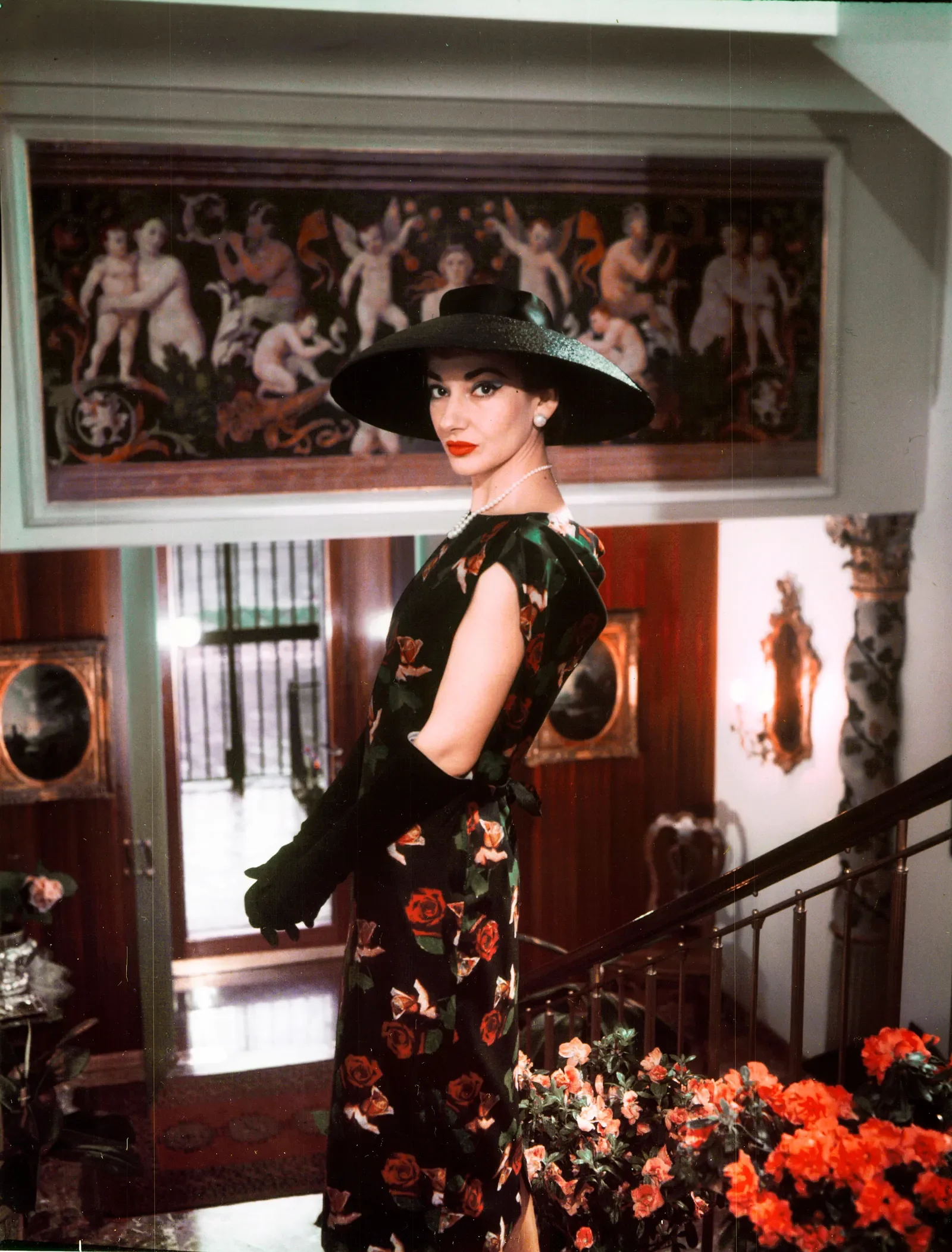 Maria Callas com vestido estampado e chapéu, em ambiente sofisticado.