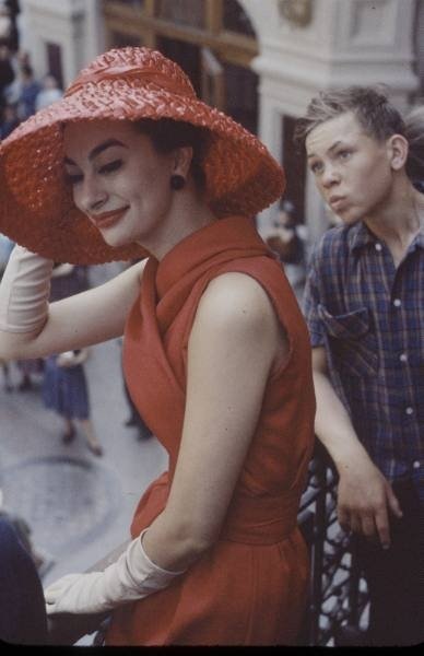 Maria Callas de vestido e chapéu vermelho em uma varanda, acompanhada por rapaz.
