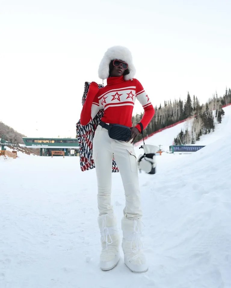 Pessoa em roupa de neve com suéter vermelho decorado, óculos de sol e botas estilo moon boot em pista de esqui.