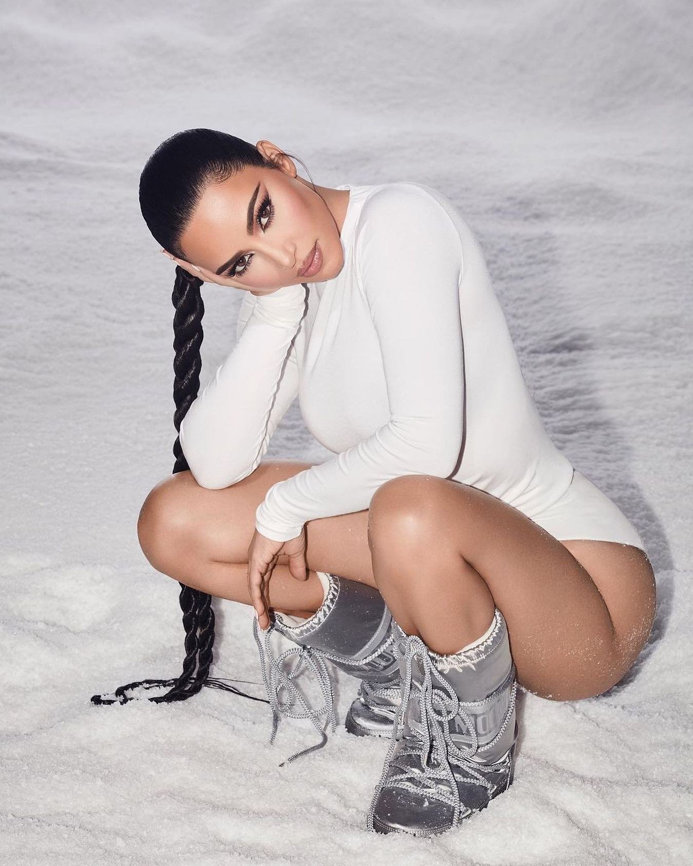 Mulher com roupa branca e botas moon boot prateadas agachada na neve, com cabelo trançado.