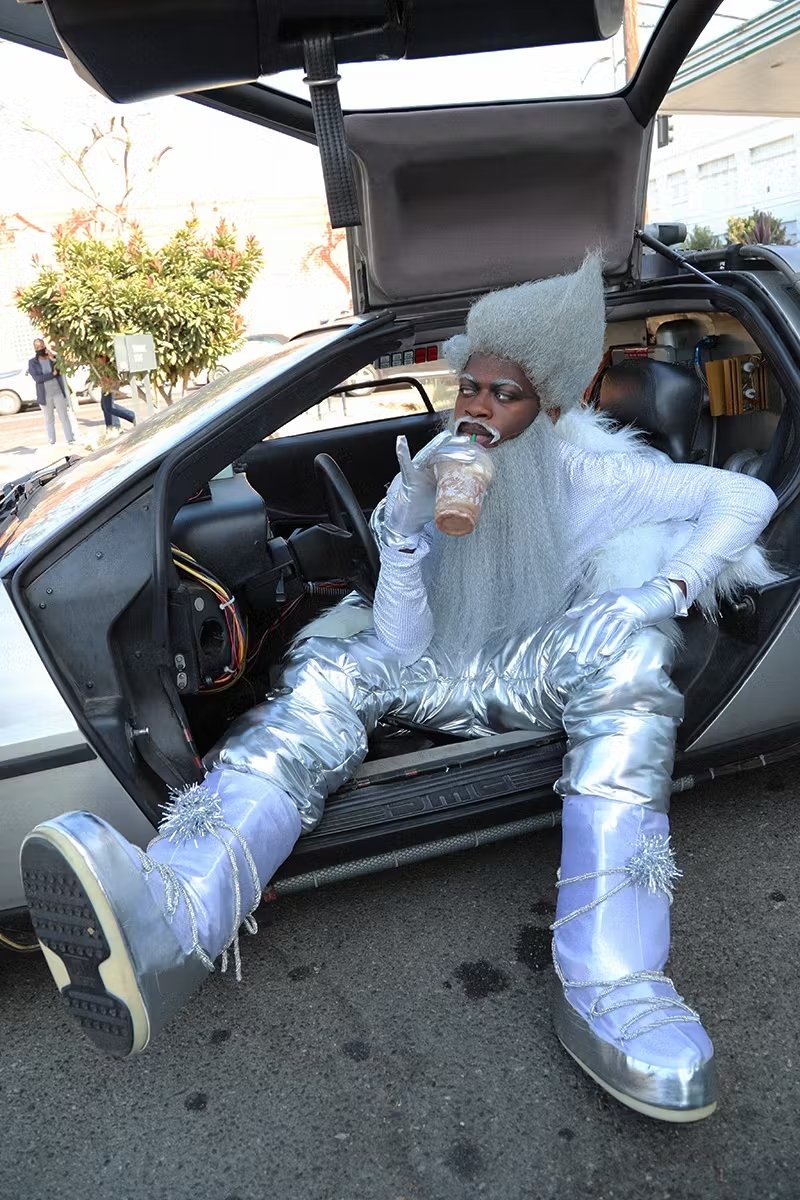 Pessoa com traje prateado e botas estilo moon boot, em um carro com porta asa de gaivota, bebe algo gelado.