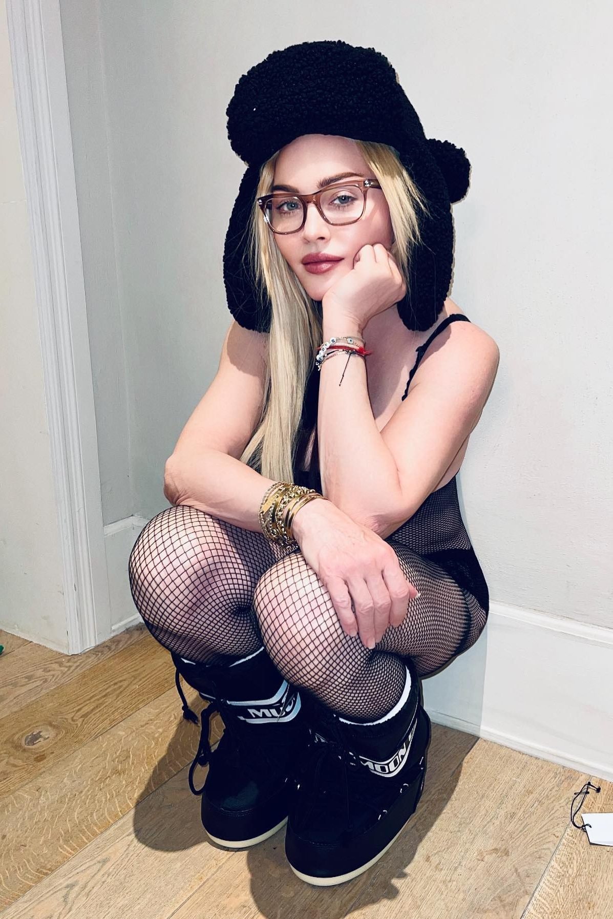 Pessoa usando chapéu peludo preto, óculos e botas Moon Boot pretas, agachada em um chão de madeira.