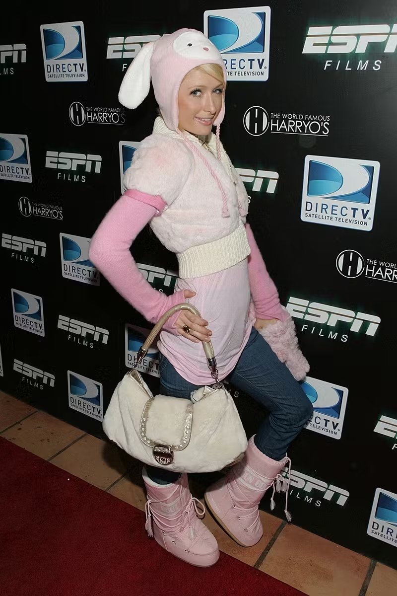 Pessoa em roupa rosa com capuz de coelho, carregando bolsa branca. Usa moon boots rosas, posando em tapete vermelho.