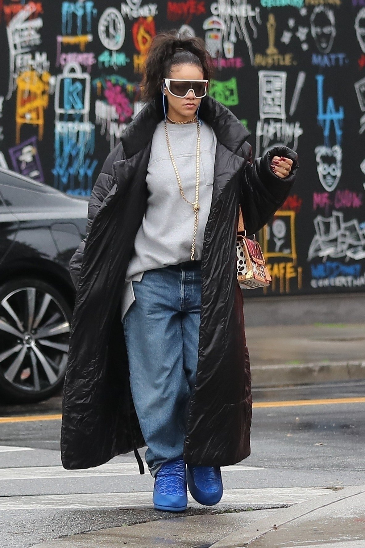 Pessoa com casaco longo preto, jeans e botas azuis estilo moon boot, em frente a mural grafitado.