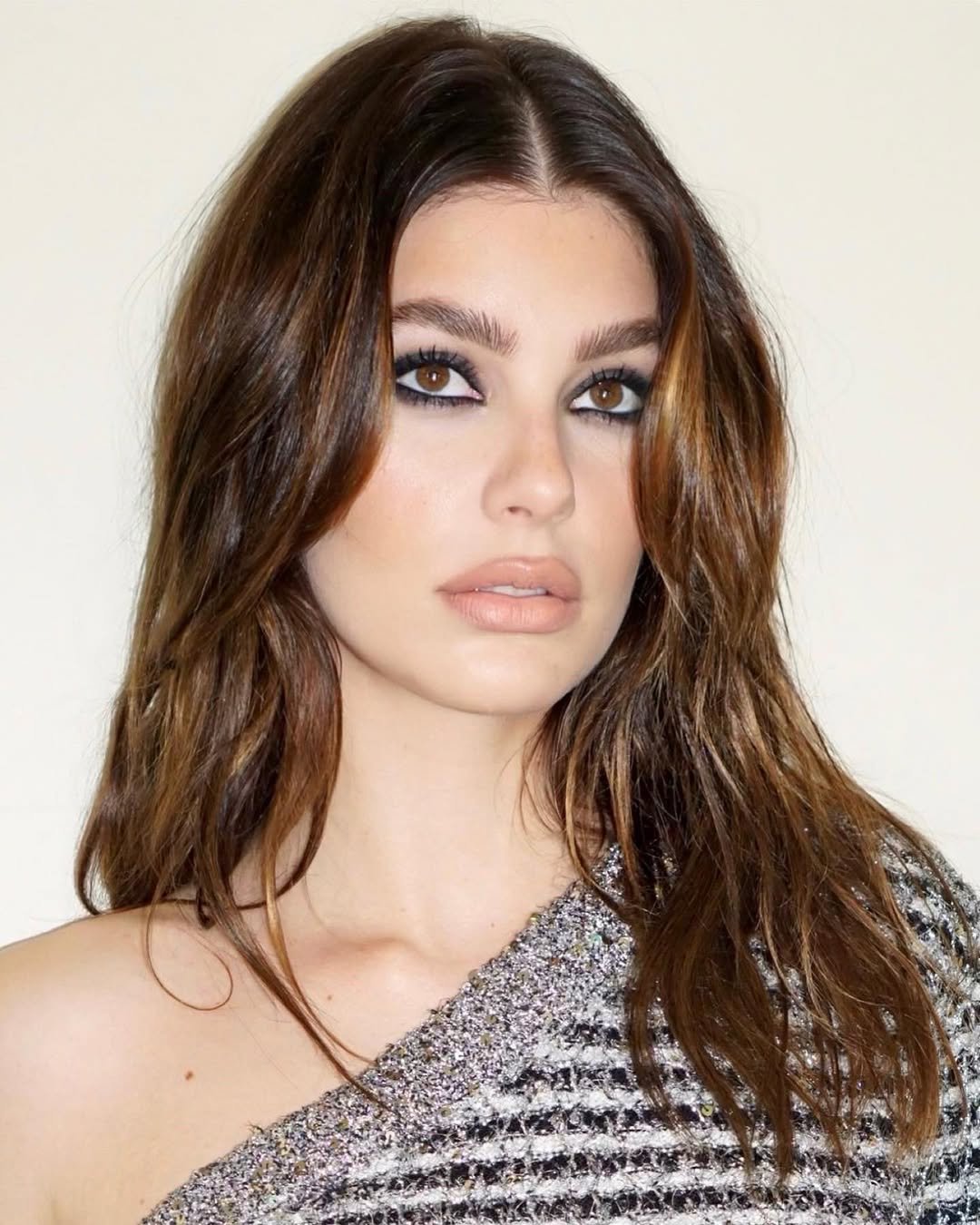 Mulher com cabelo castanho claro e olhos destacados por maquiagem, vestindo top cintilante. Técnica morena iluminada avelã aplicada.