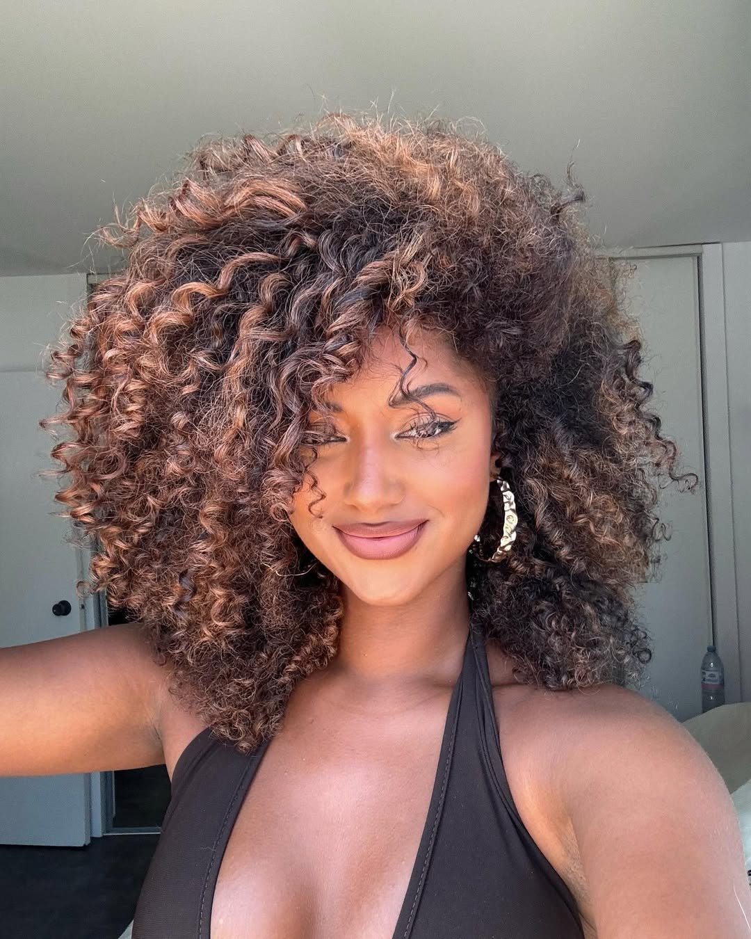 Mulher sorridente com cabelo cacheado morena iluminada avelã, usando blusa preta e brincos grandes.