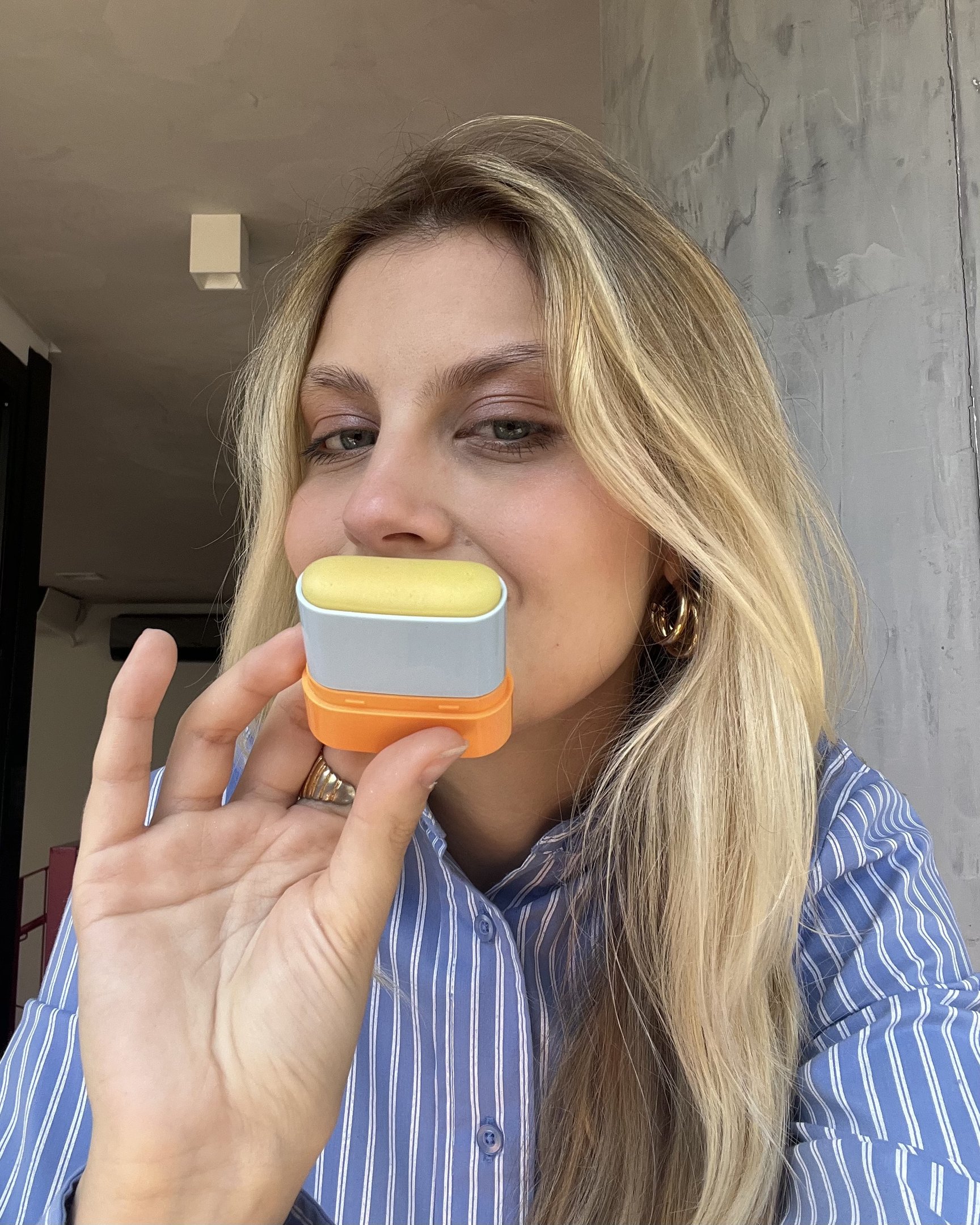Pessoa segura um protetor solar ISDIN em formato compacto com tampa amarela e base laranja.
