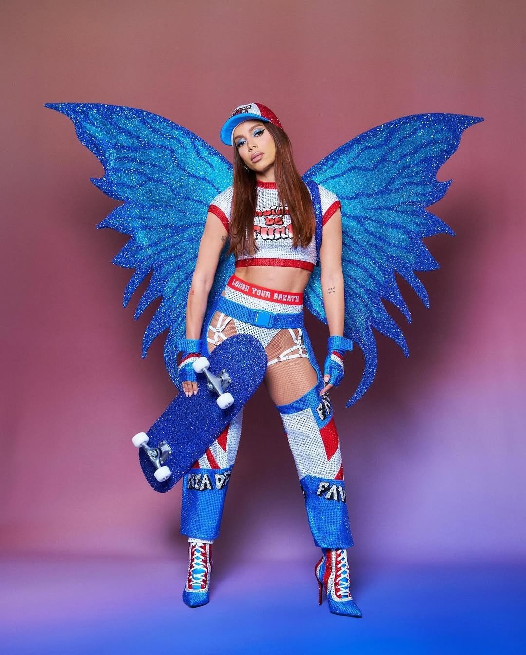 Anitta com fantasia azul e vermelha, asas de borboleta e segurando um skate azul, em pose confiante.
