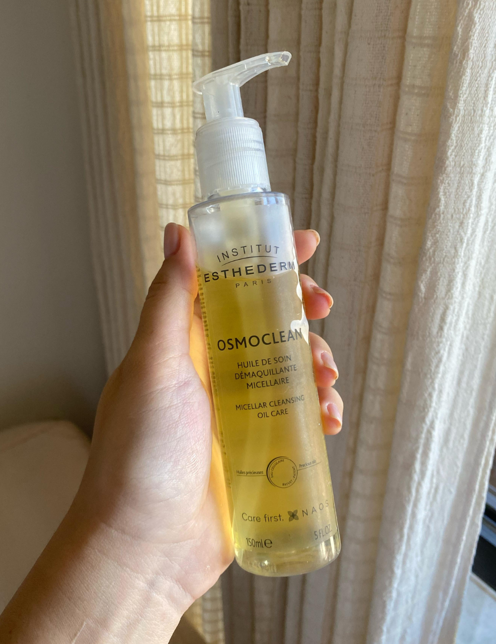 Frasco de óleo micelar para limpeza de pele, segurado por uma mão, em ambiente iluminado.