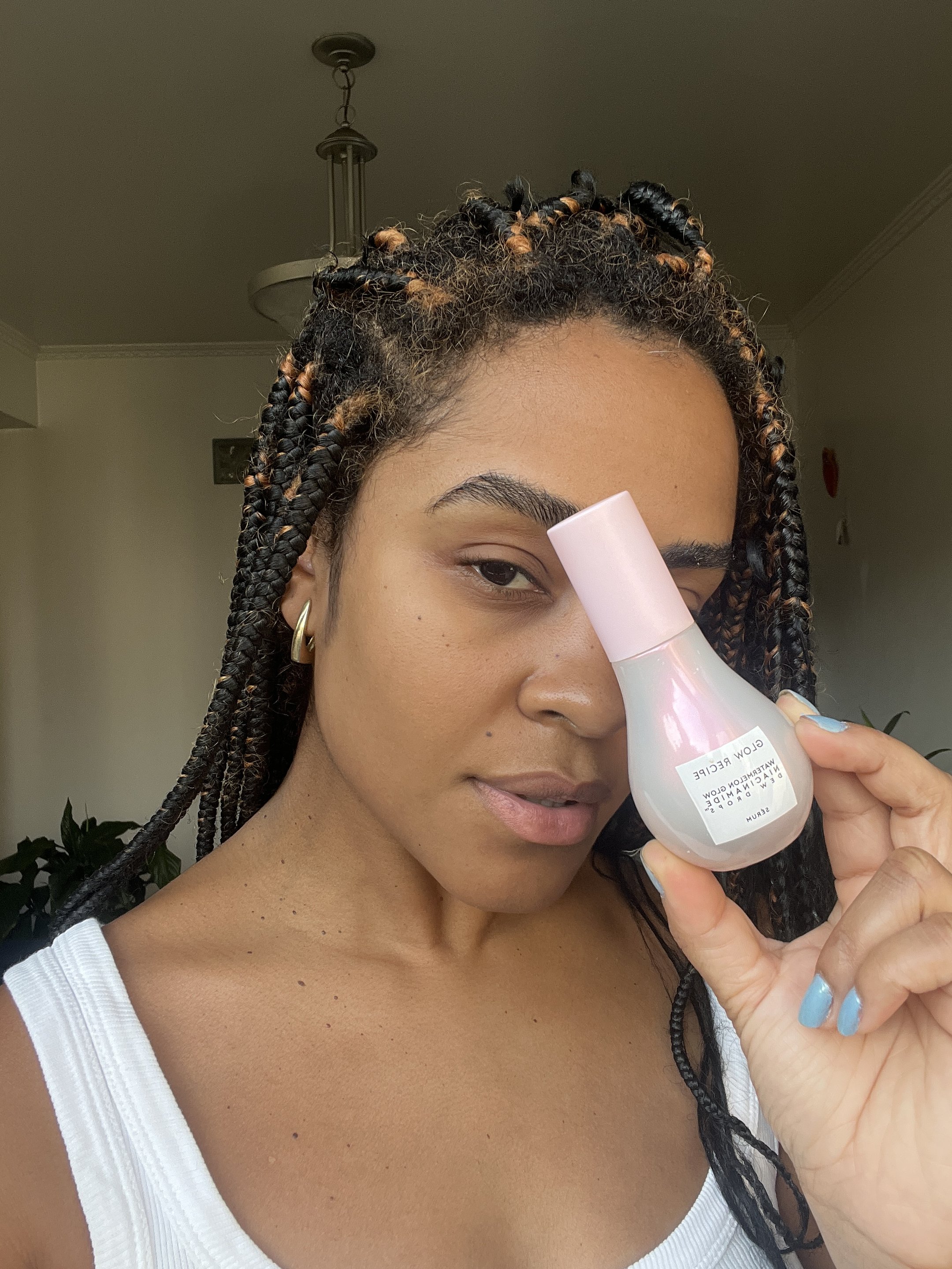Pessoa segurando um produto da Glow Recipe próximo ao rosto, com frasco rosa e ambiente iluminado ao fundo.