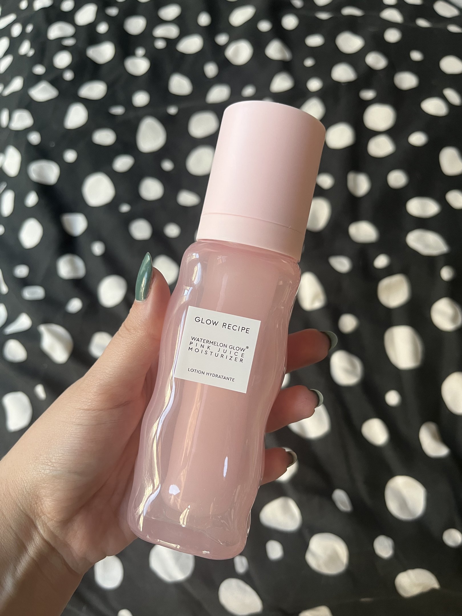 Loção hidratante produtos da Glow Recipe em embalagem rosa, segurada por uma mão sobre fundo preto com bolinhas brancas.