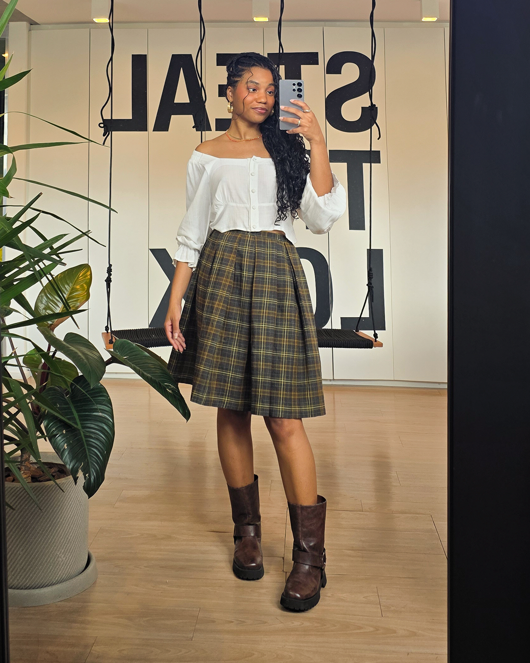 Saia xadrez combinada com blusa branca e botas marrons refletida em espelho, com plantas ao redor e janela ao fundo.