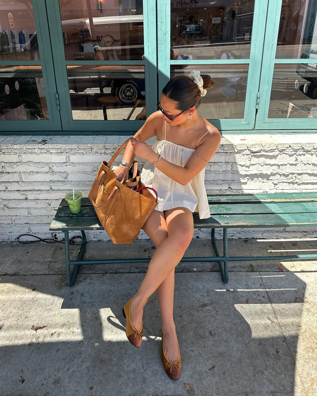 Pessoa sentada em banco, vestindo vestido claro e sapatos casuais, mexendo numa bolsa. Bebida ao lado.