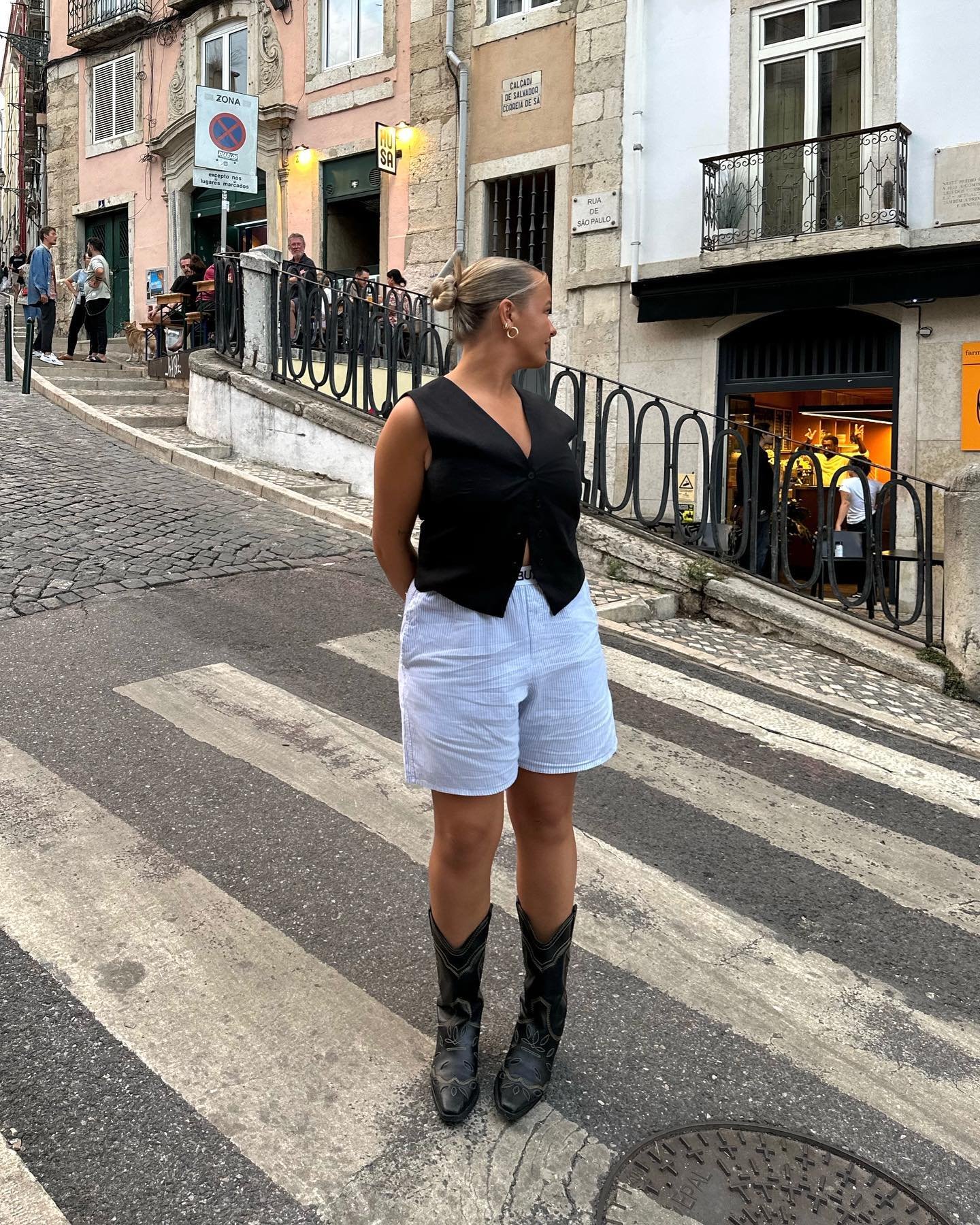 Pessoa com botas de sapato retrô, shorts claros e blusa preta na faixa de pedestres em rua urbana.