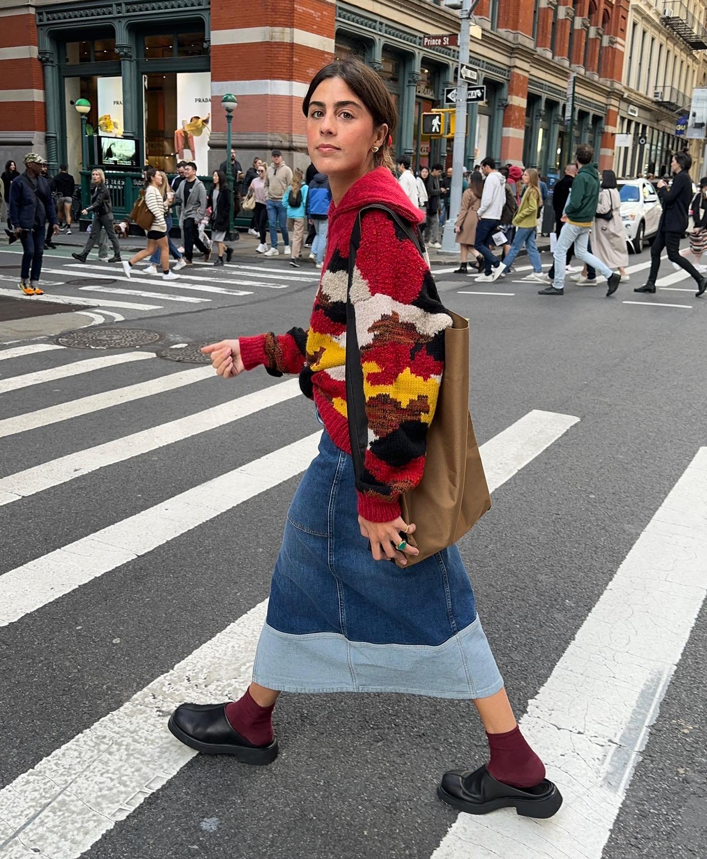 Pessoa atravessa a rua usando suéter colorido, saia jeans e sapatos retrô. Trânsito urbano ao fundo.