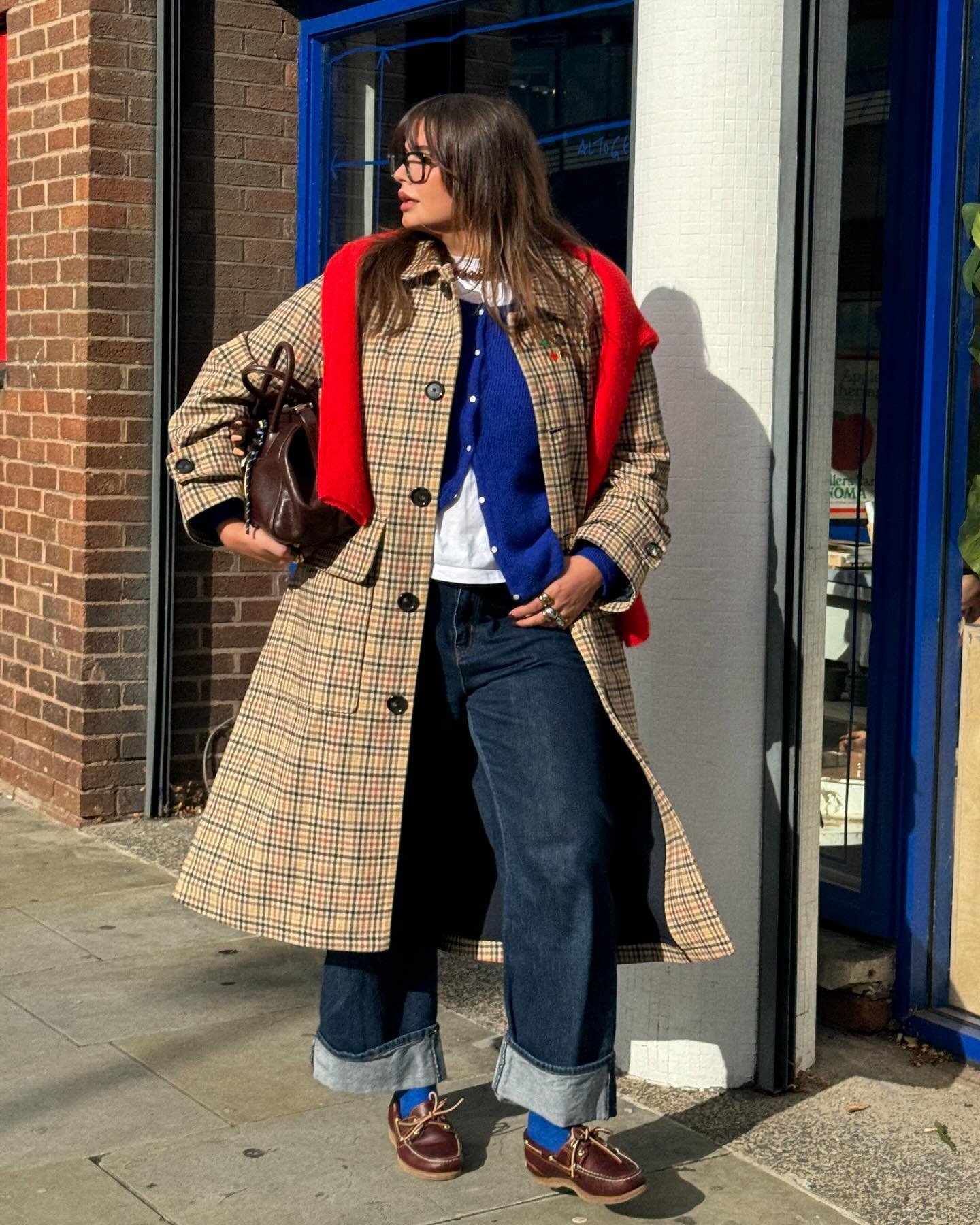 Casaco xadrez longo sobre jeans e blusa azul com suéter vermelho. Estilo urbano segue principais tendências de 2025.