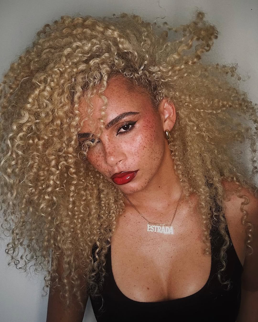 Cabelo cacheado volumoso e loiro, Batom vermelho para iniciantes brilhante, olhar marcante, pele com sardas. Colar com nome.