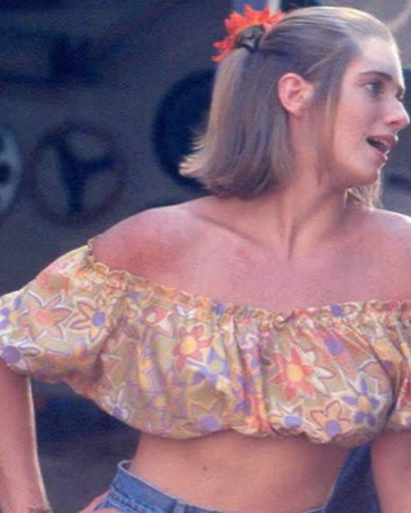 Mulher com cabelo preso usa blusa colorida ombro a ombro, estilo típico de personagens em novelas.