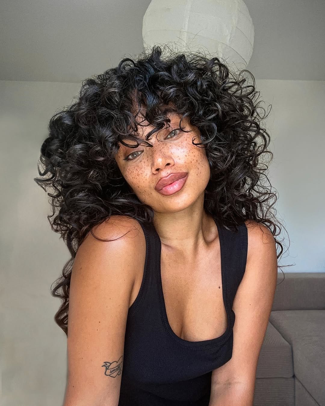 Mulher com cabelos cacheados volumosos e uma regata preta, destacando estilo natural do Guia do cabelo cacheado.