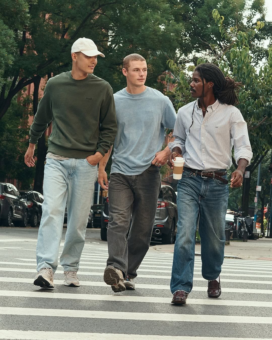 Três homens conversam enquanto atravessam uma rua arborizada usando jeans oversized 
