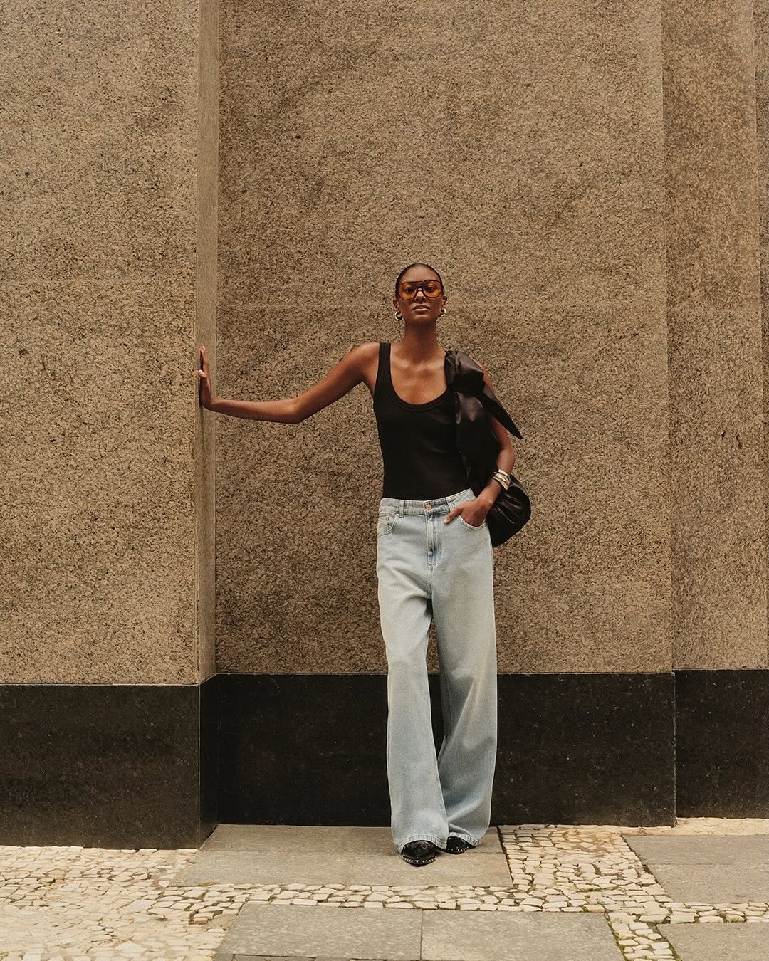 Pessoa com look minimalista elegante, usa regata preta e calça jeans larga, encostada em parede de pedra.