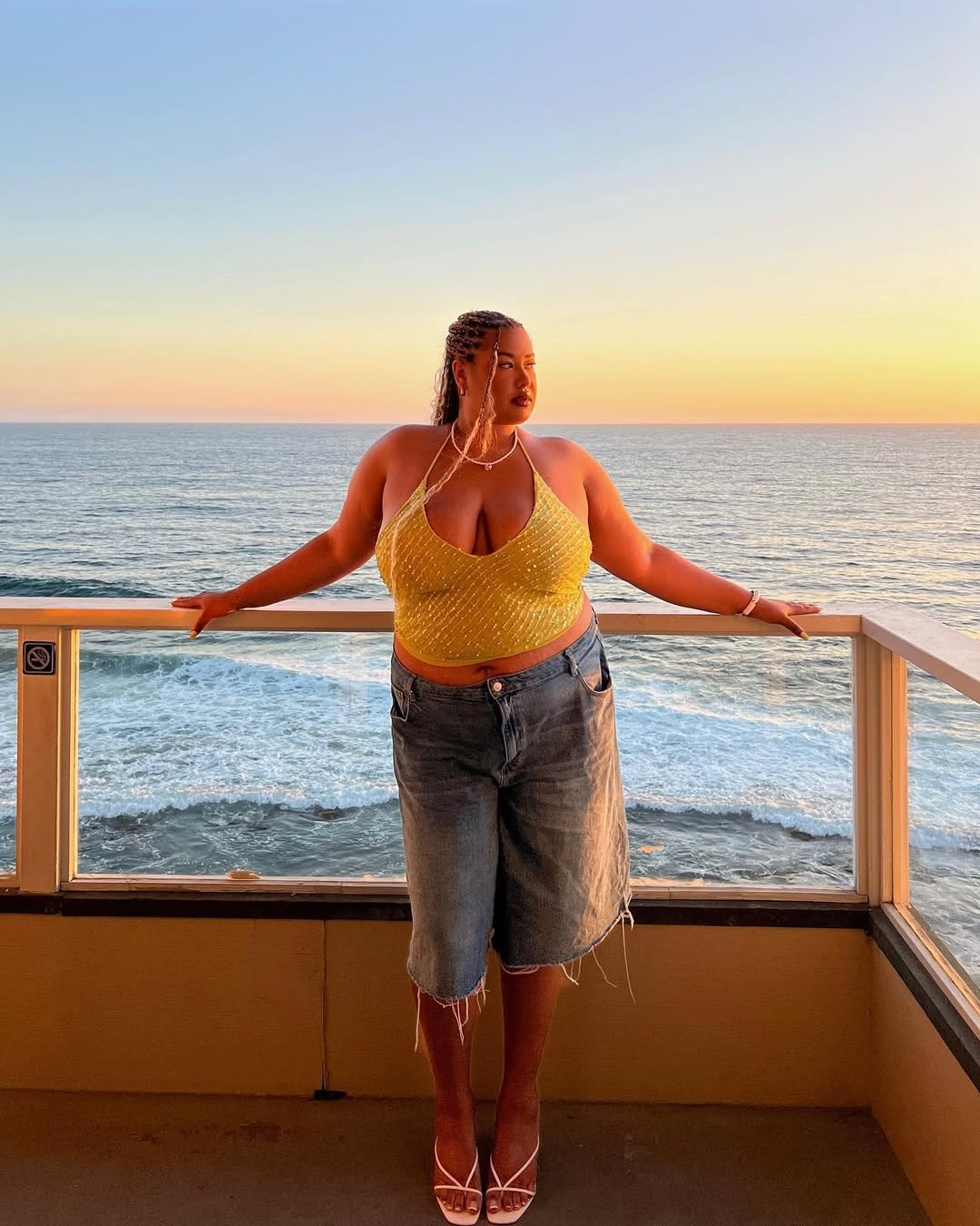 Pessoa em varanda com vista para o mar ao pôr do sol. Vestindo blusa amarela e bermuda jeans. Suco de aloe vera na mente.