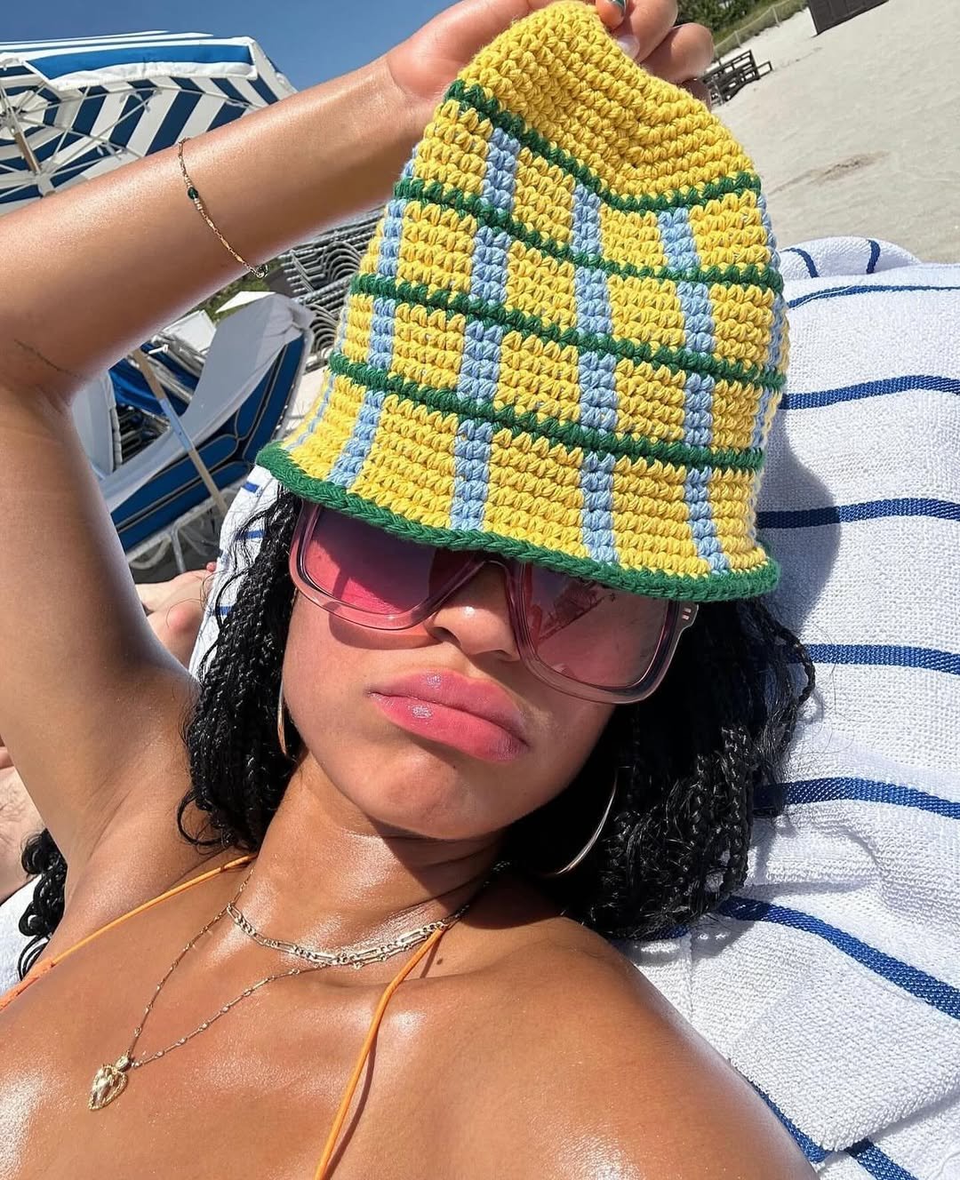 Pessoa deitada na praia com chapéu de crochê xadrez amarelo e óculos de sol rosas. O conceito de relaxamento combina com suco de aloe vera.