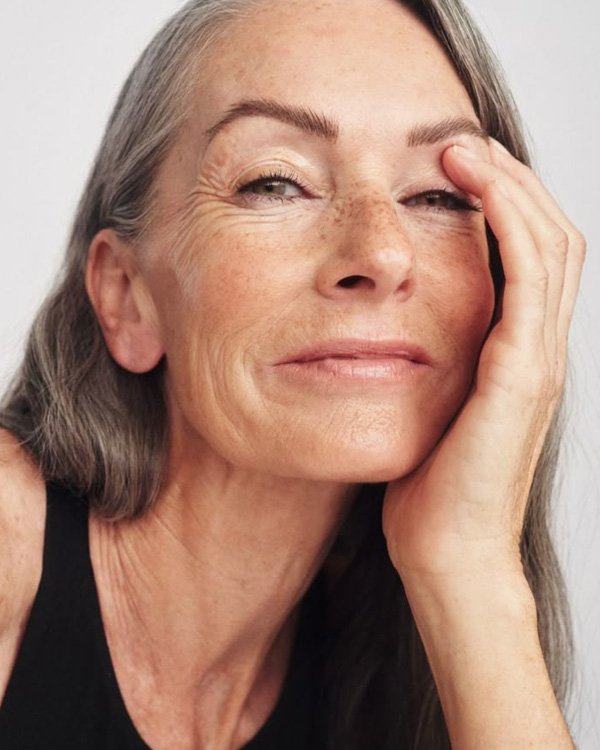 Mulher madura com expressão confiante, pele iluminada, possivelmente usando creme noturno, apoiando o rosto com a mão.