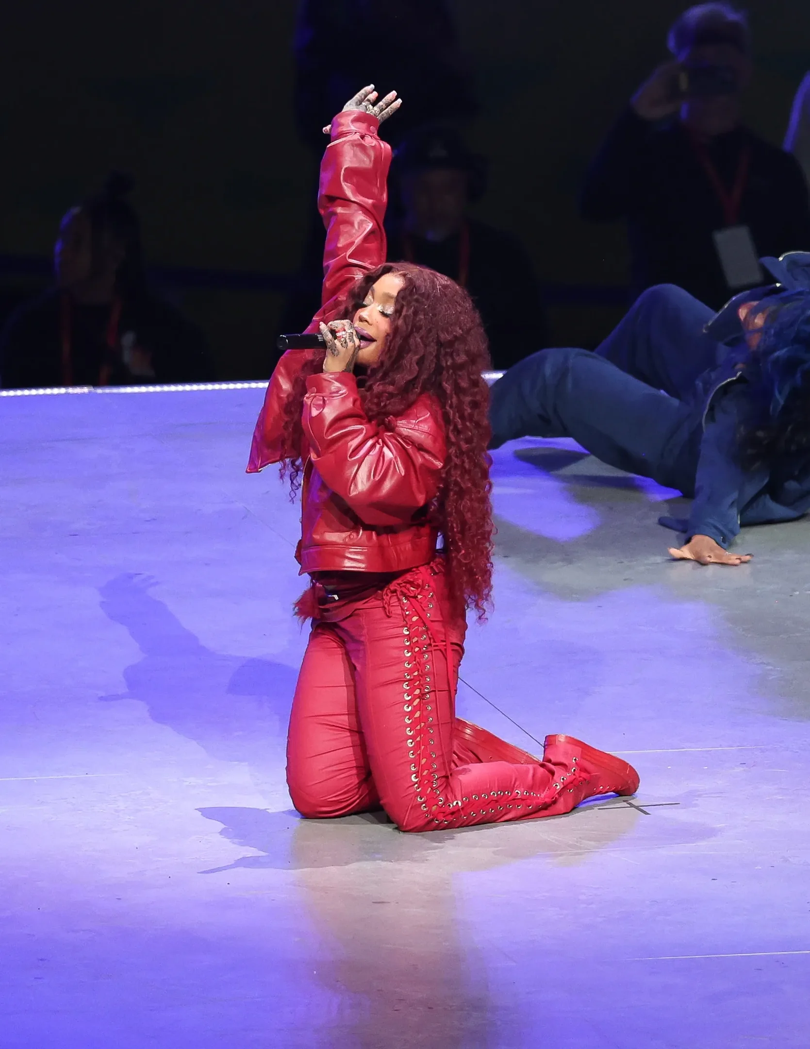 SZA está ajoelhada no palco com traje vermelho, cantando com entusiasmo.