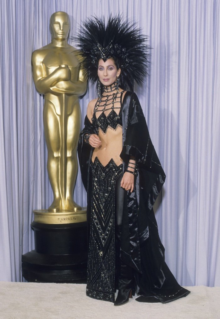 Celebridade com traje ousado e glamouroso ao lado de uma estátua dourada, exemplificando looks do tapete vermelho.