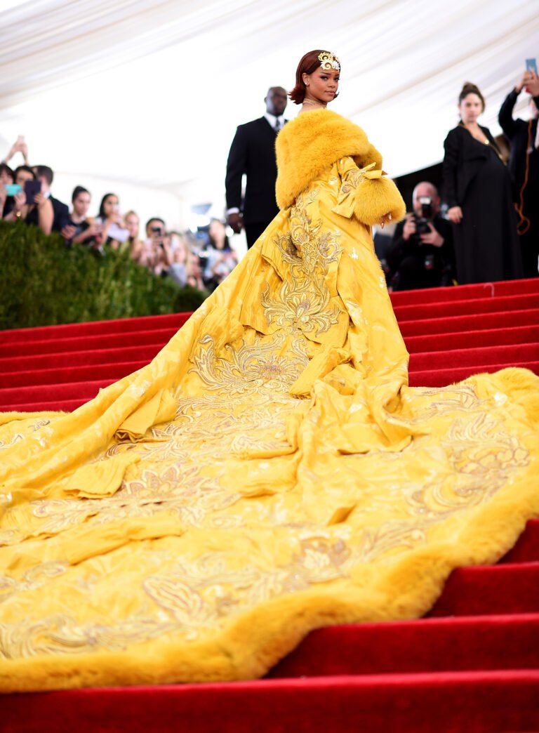 Vestido amarelo com cauda longa em evento sofisticado nos looks do tapete vermelho, cercado por fotógrafos e público.