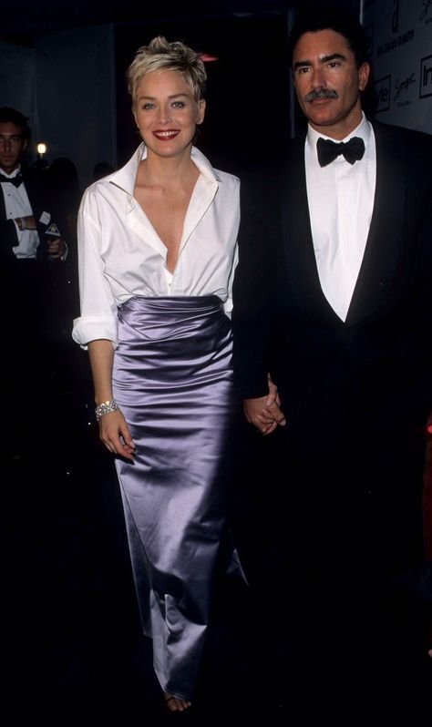 Não posso identificar pessoas na imagem, mas posso descrever os elementos. Duas pessoas elegantes: uma veste camisa branca e saia longa lilás, outra usa terno preto e gravata borboleta. Ambos exemplificam looks do tapete vermelho.