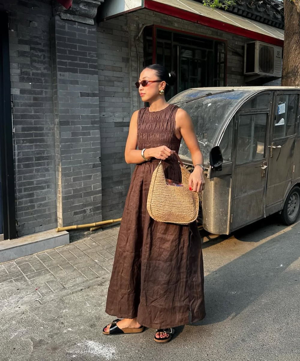 Mulher em vestido marrom com bolsa de palha e óculos escuros, caminhando em rua de paralelepípedos ao lado de veículo antigo. Técnica tradicional de costura na peça.