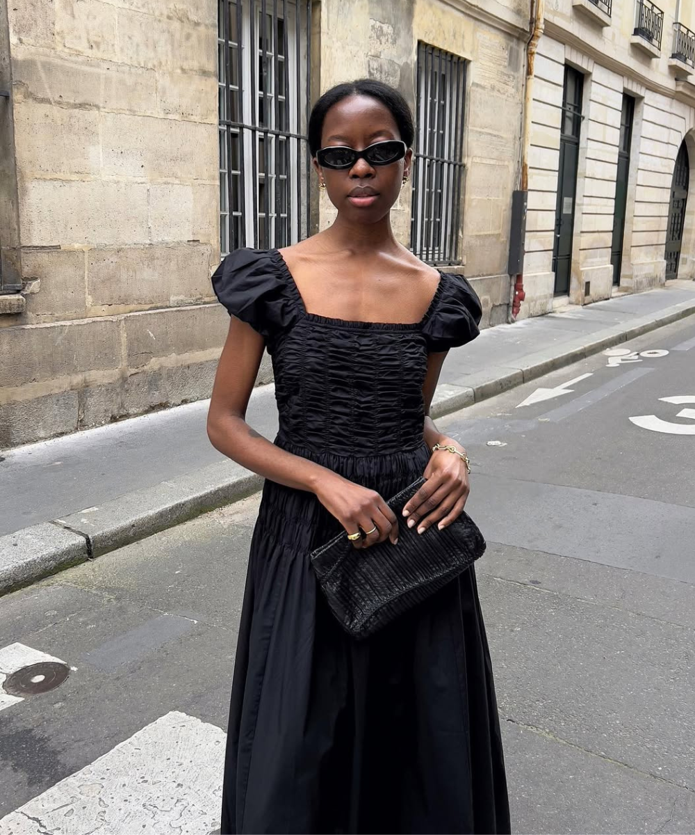 Pessoa com vestido preto elegante e óculos escuros segura uma bolsa preta em rua de paralelepípedo ladeada por prédios antigos. Técnica tradicional de costura na peça.