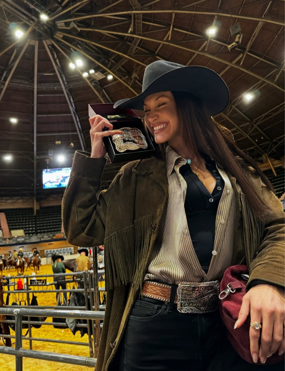 Mulher sorridente em evento de montaria, usando chapéu e cinto com fivela grande. Estilo remete a tendências vintage.