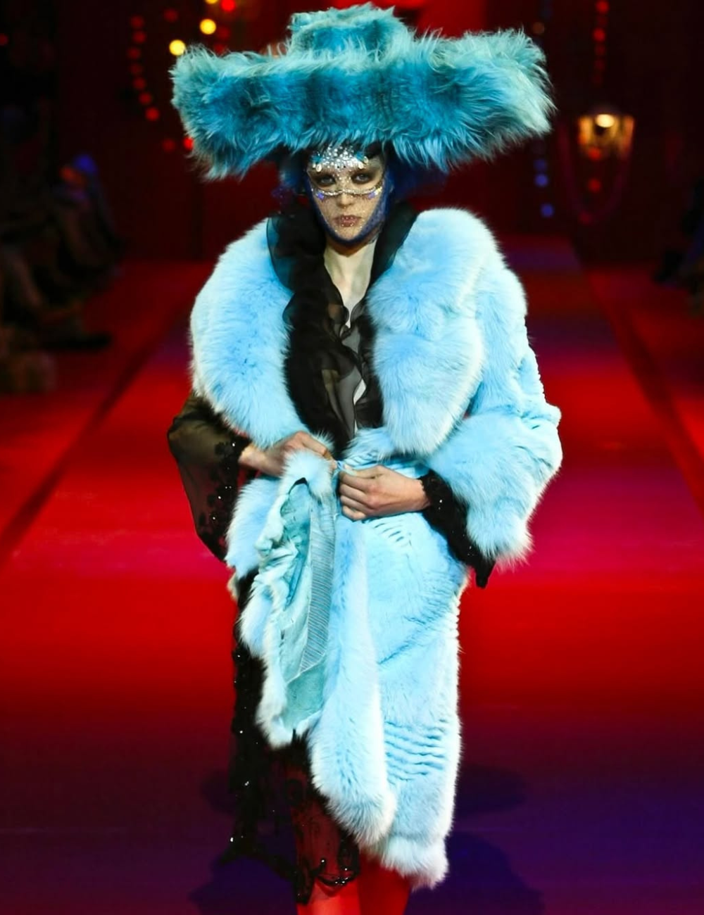 Modelo com casaco e chapéu azul peludo extravagantes, maquiagem com brilho, em passarela com tendências vintage.