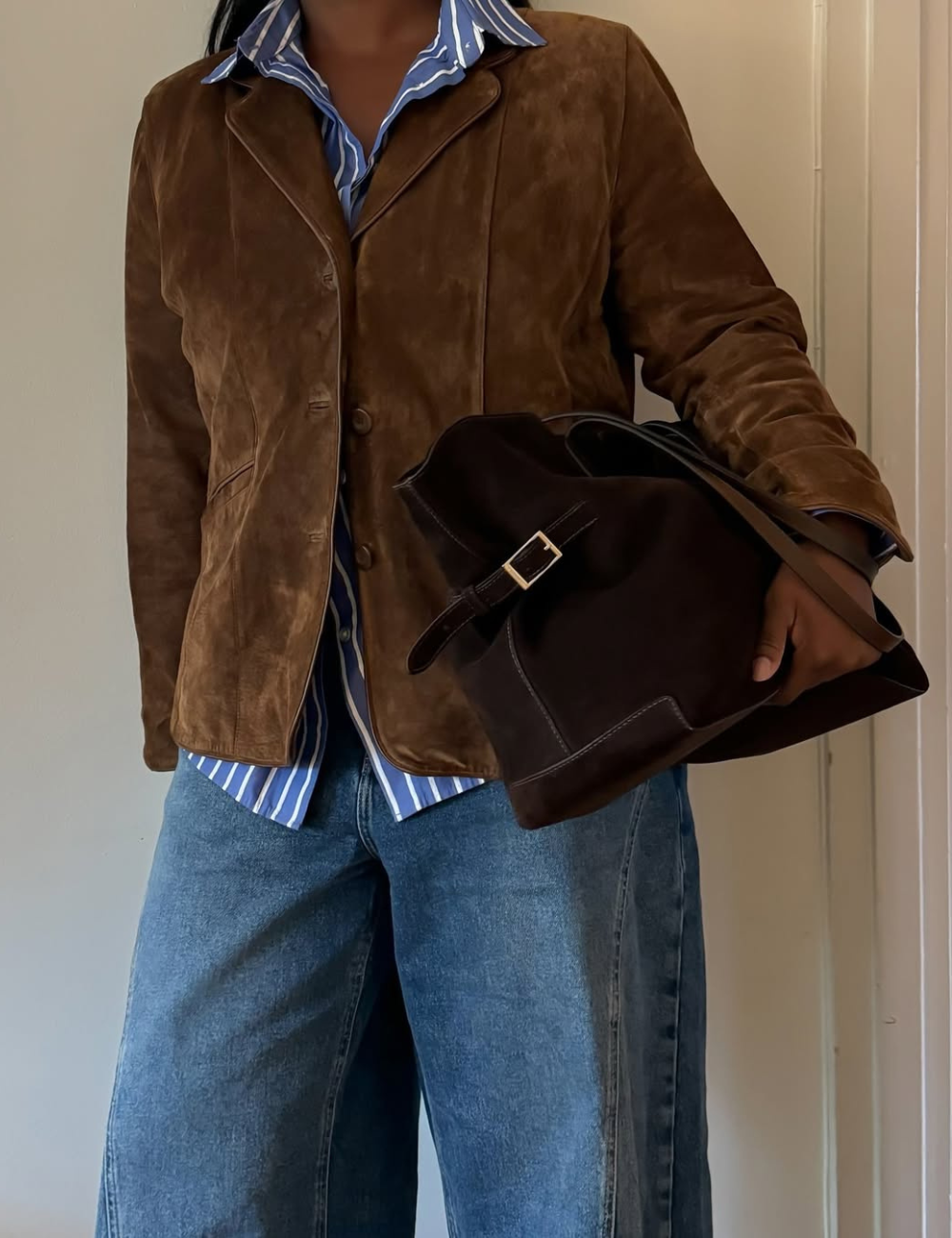 Casaco de camurça marrom, camisa listrada e calça jeans remetem às tendências vintage. Pessoa segura bolsa escura.