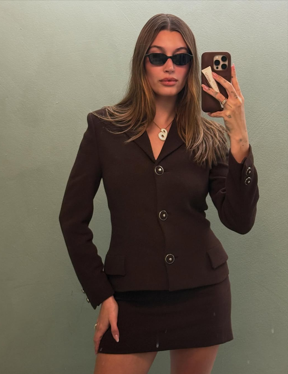 Mulher usa blazer e saia marrom com óculos escuros, refletindo tendências vintage.