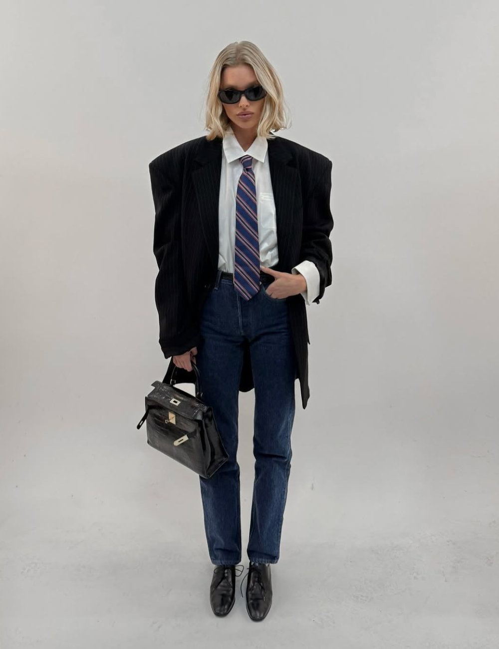 Pessoa com óculos escuros, blazer largo, gravata listrada, jeans e bolsa preta, exibindo tendências vintage.